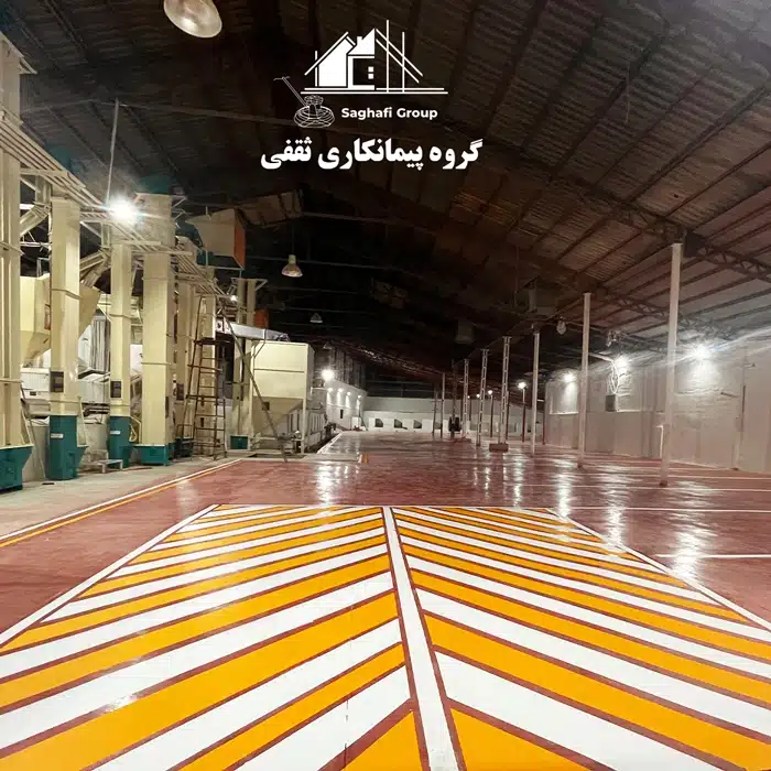 تصویر داخلی یک شالیکوبی با کفپوش صنعتی قرمز رنگ و خط‌کشی‌های ایمنی زرد و سفید. ماشین‌آلات شالیکوبی در سمت چپ و فضای خالی انبار در سمت راست قابل مشاهده است.